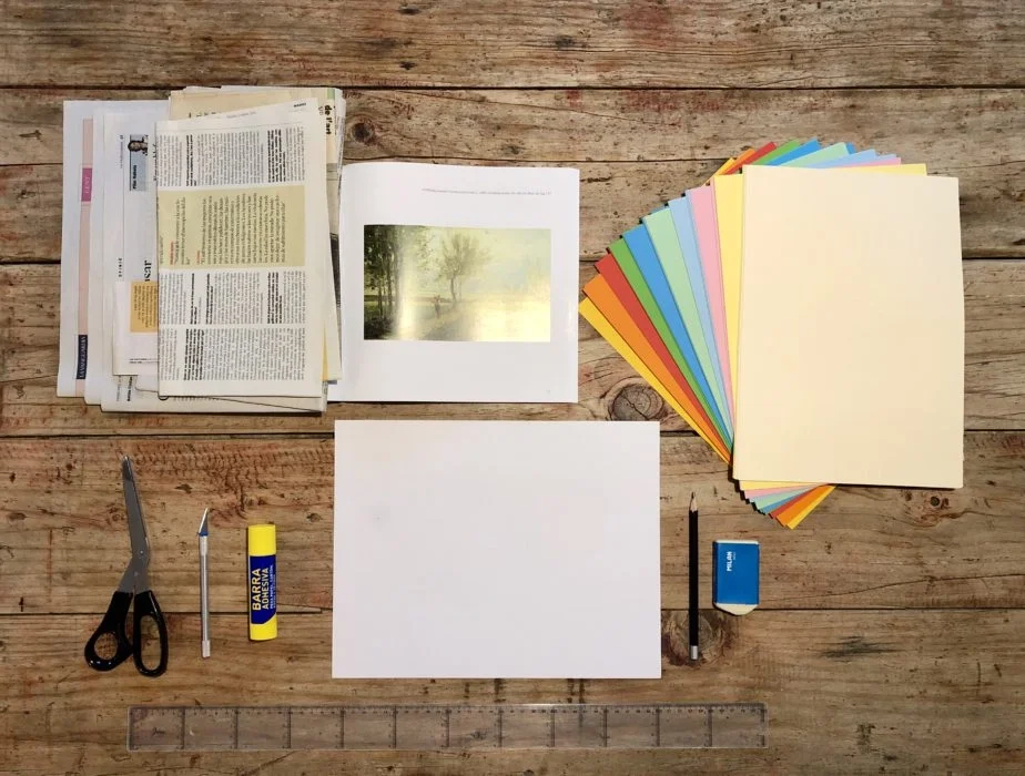 Imatge amb fulls de papers de colors a la dreta, un full en blanc al centre, un llapis i una goma a la dreta del full en blanc, unes tisores i un cuter a l'esquerra del full en blanc i uns diaris i fotografies retallables a la part superior esquerra